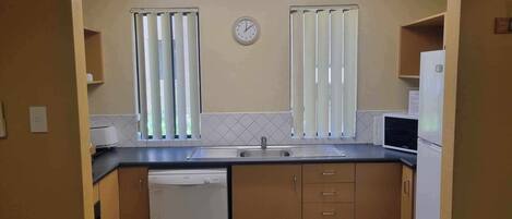 A compact kitchen with a dishwasher, a microwave, and a refrigerator. There are two windows with blinds, a clock on the wall, and a tiled backsplash.