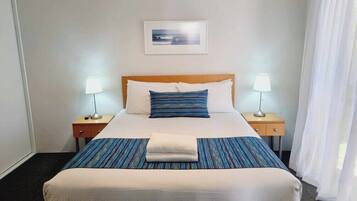Ein ordentlich bezogenes Bett mit blau-weiß gestreifter Tagesdecke, einem weißen Kissen und einem gefalteten weißen Handtuch. Darüber an der Wand hängt ein gerahmtes Bild, und auf beiden Seiten des Bettes stehen Nachttische mit Lampen.