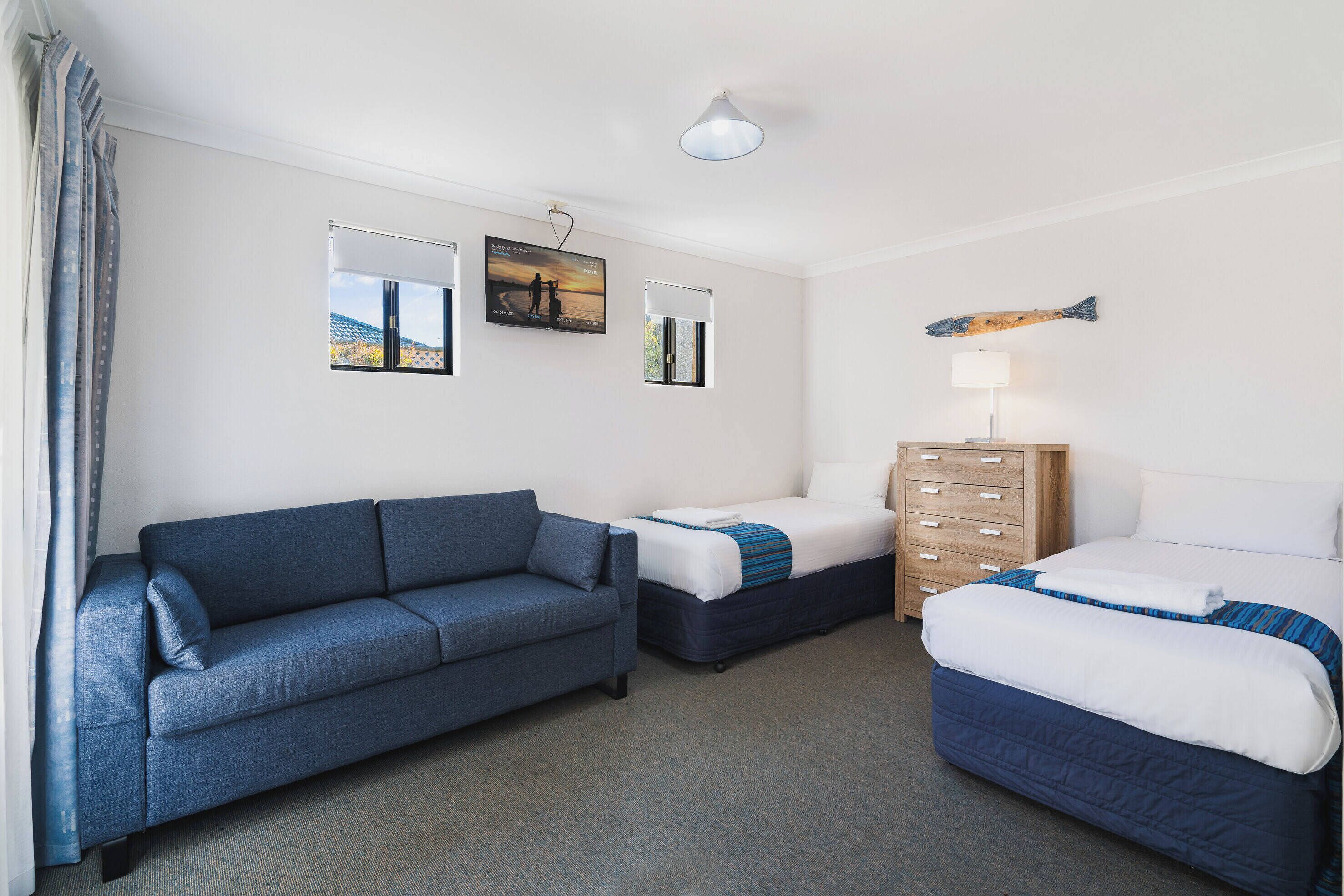 Une chambre d’hôtel avec un canapé bleu, un lit recouvert d’une courtepointe bleue et blanche, une commode en bois et deux téléviseurs fixés au mur diffusant une scène de coucher de soleil.