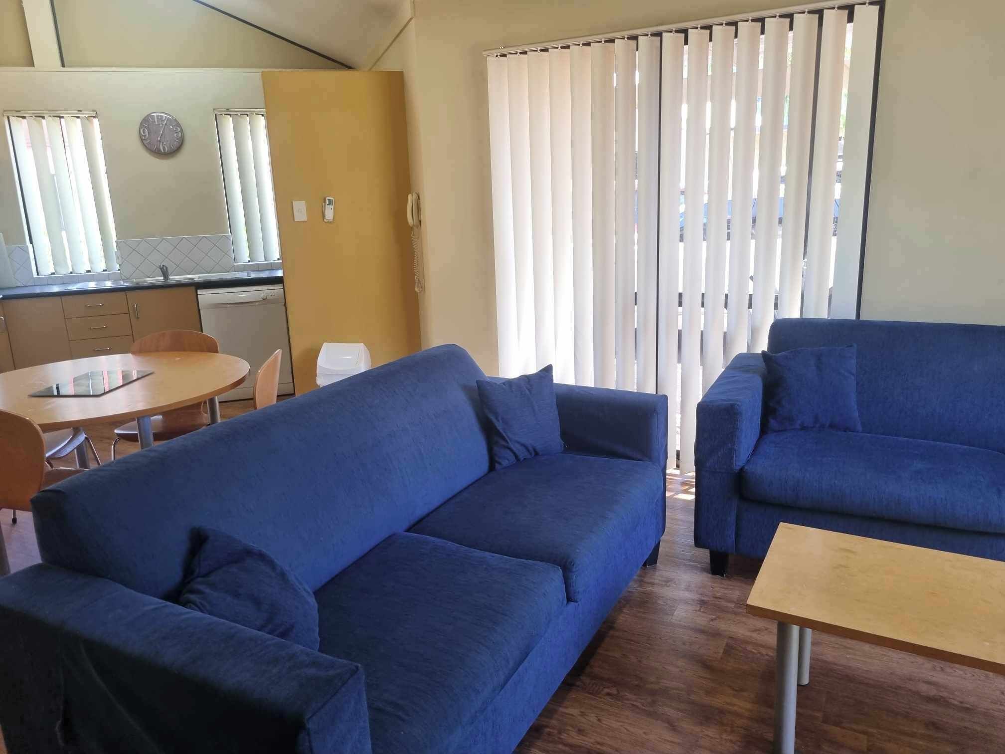 Una sala de estar con dos sofás azules, una mesa de centro de madera, y una zona de cocina con un fregadero y armarios.