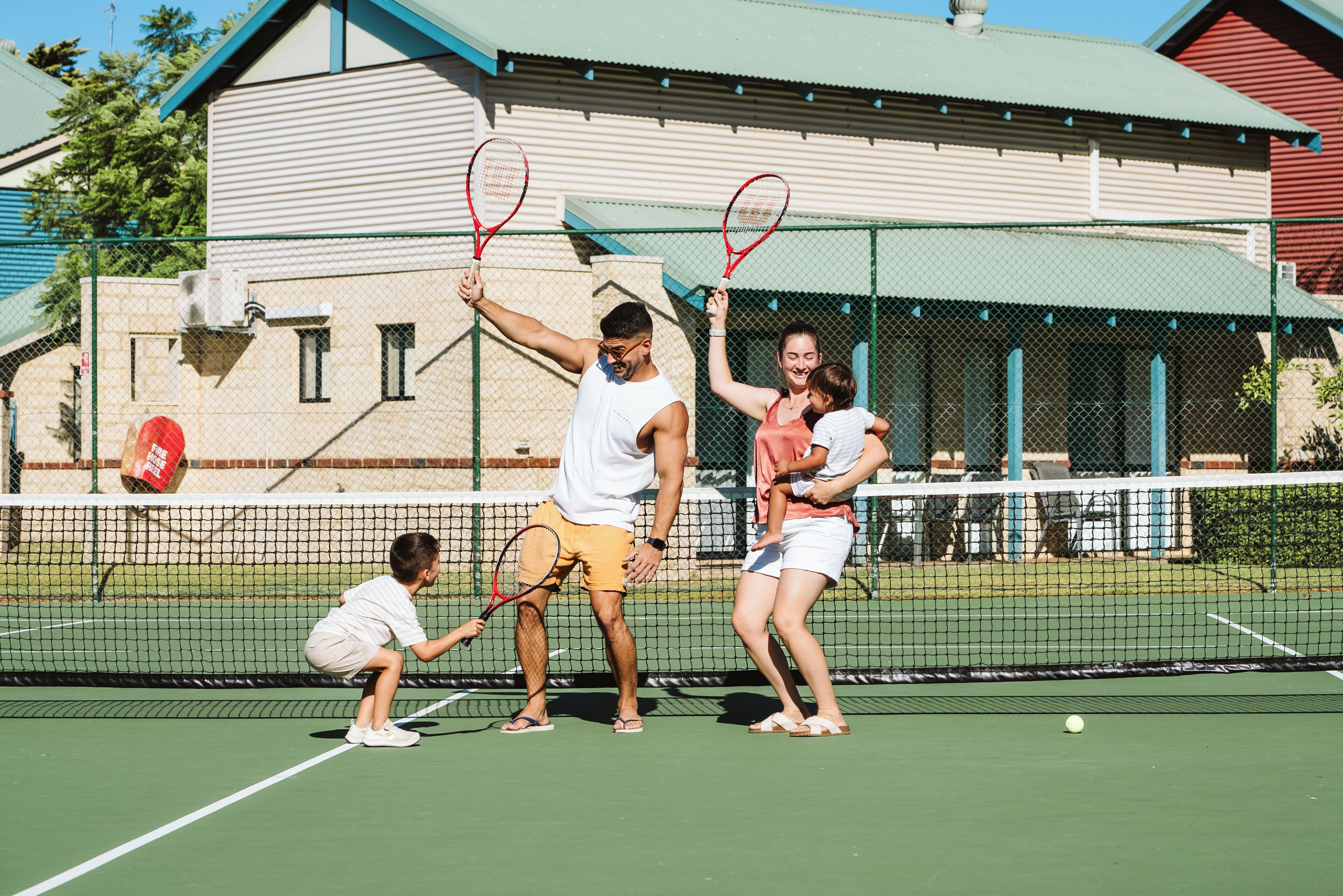 A tennis court with a net, a building with a green roof, and a fence in the background.