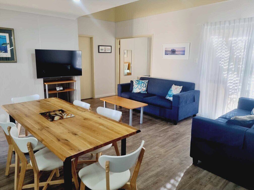 A living room with a wooden dining table, white chairs, a blue sofa, and a television.
