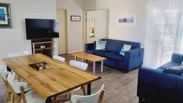 A living room with a wooden dining table, white chairs, a blue sofa, and a television.