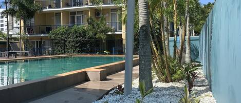 A pool area with a clear blue sky, a yellow building with balconies, and a palm tree.