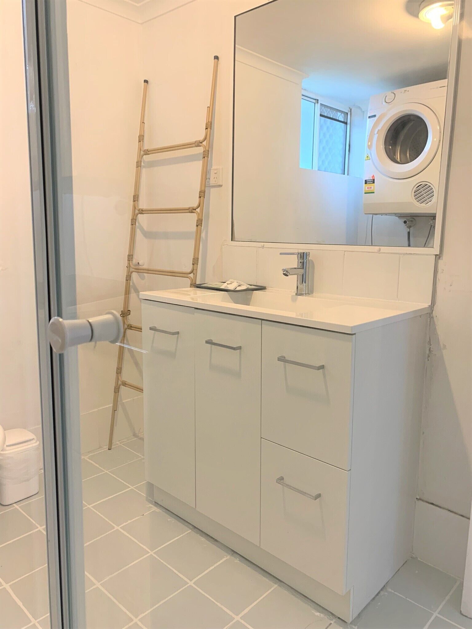 Casa de banho com cabine de duche em vidro, lavatório branco com espelho em cima, máquina de lavar roupa e uma escada encostada à parede.