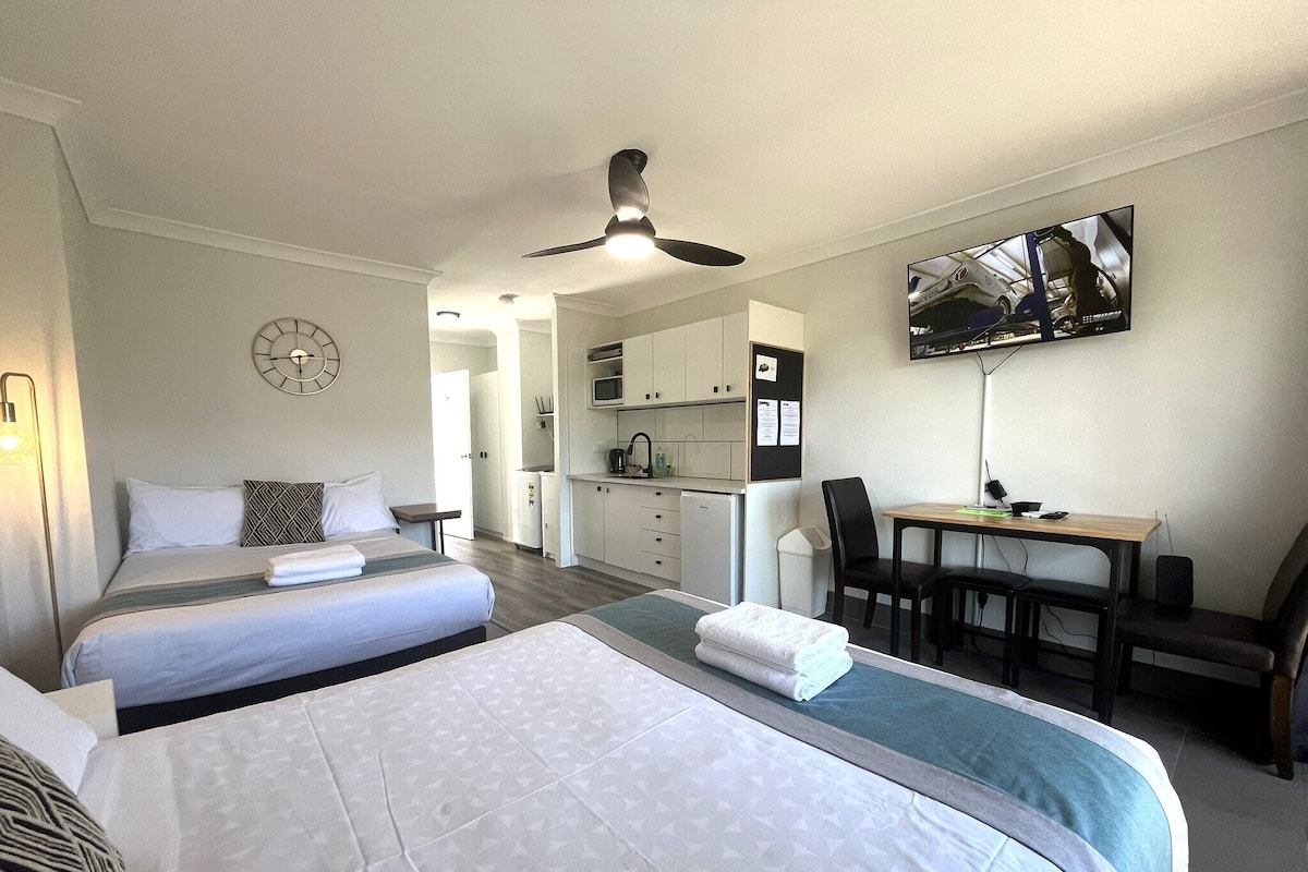 A hotel room with two beds, a desk, a chair, a TV, and a kitchenette.