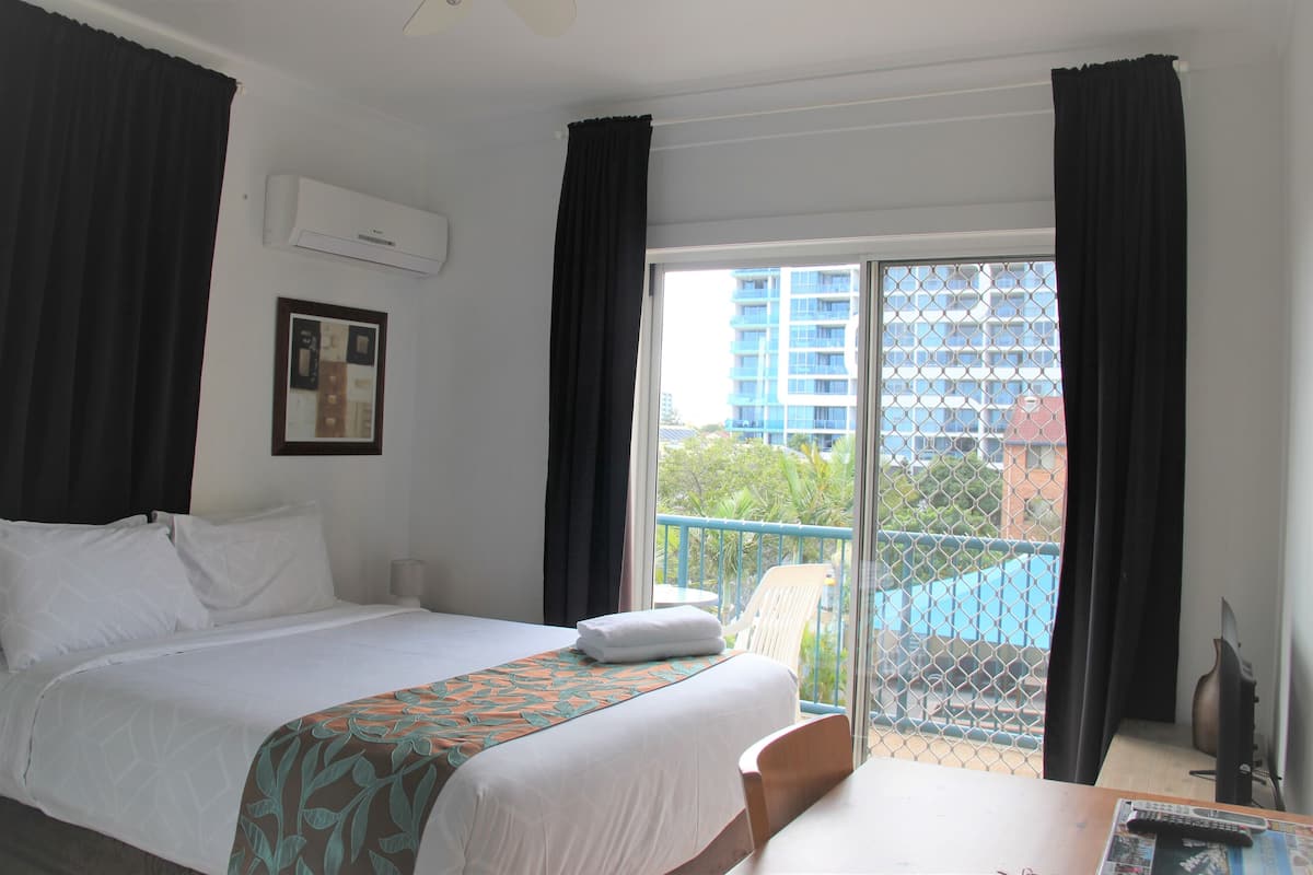A hotel room with a bed, a table, a chair, an air conditioner, and a balcony with a view of buildings and a pool.