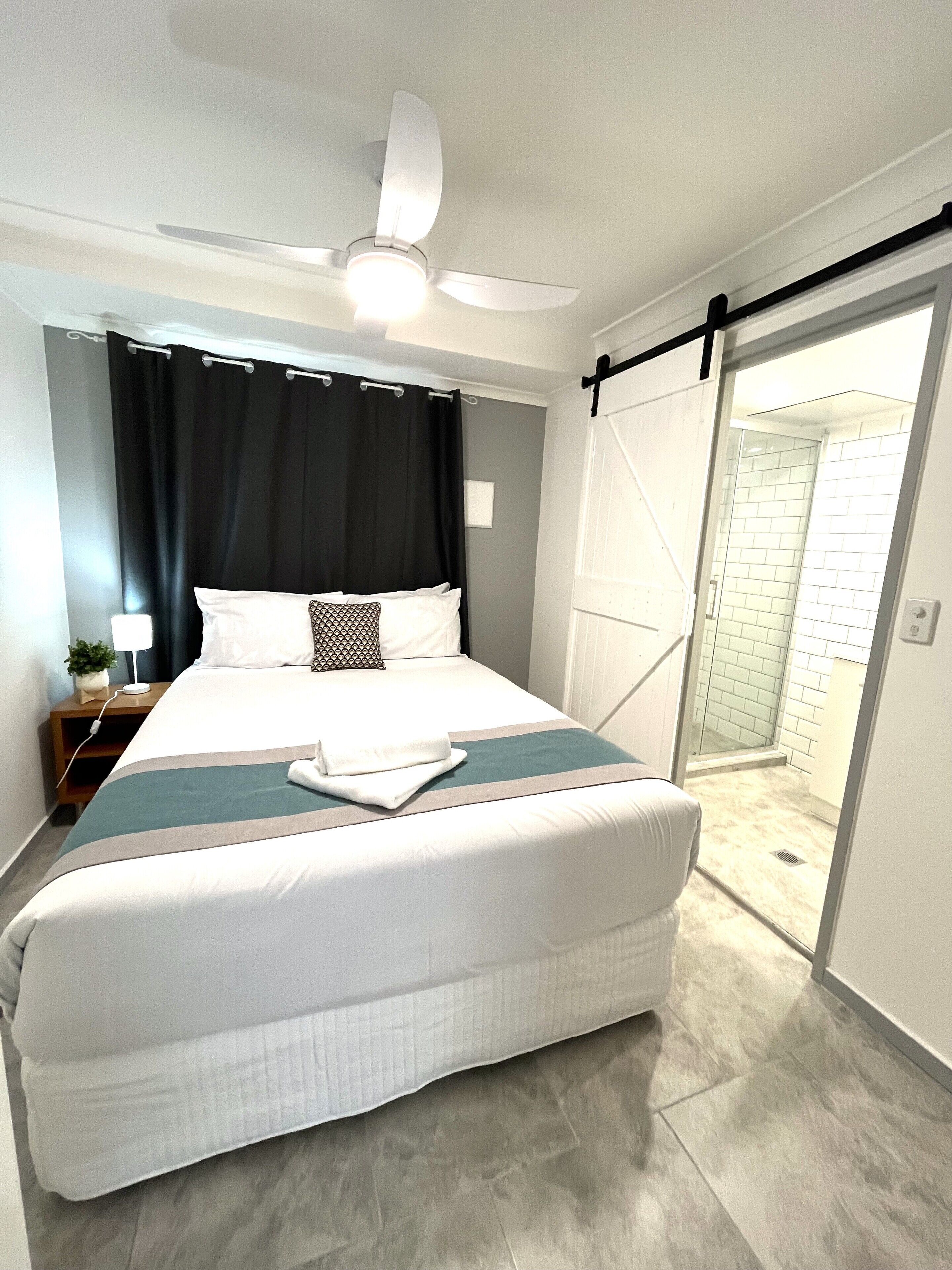 A bedroom with a bed, a ceiling fan, a nightstand with a lamp, and a view of a bathroom with a shower.