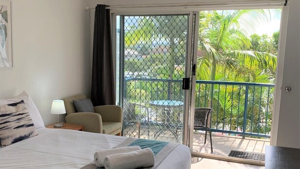 A hotel room with a bed, a desk, a chair, a balcony with a view of palm trees, and a ceiling fan. - Broadwater Keys (Labrador)