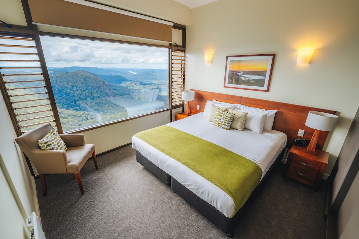 Habitación de hotel con una cama grande, una silla, una mesita de noche y vistas a un paisaje montañoso a través de un amplio ventanal.