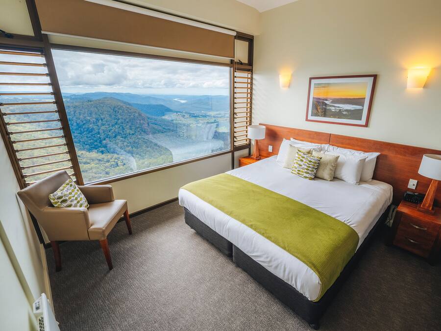 A hotel room with a large bed, a chair, a nightstand, and a view of a mountainous landscape through a large window.
