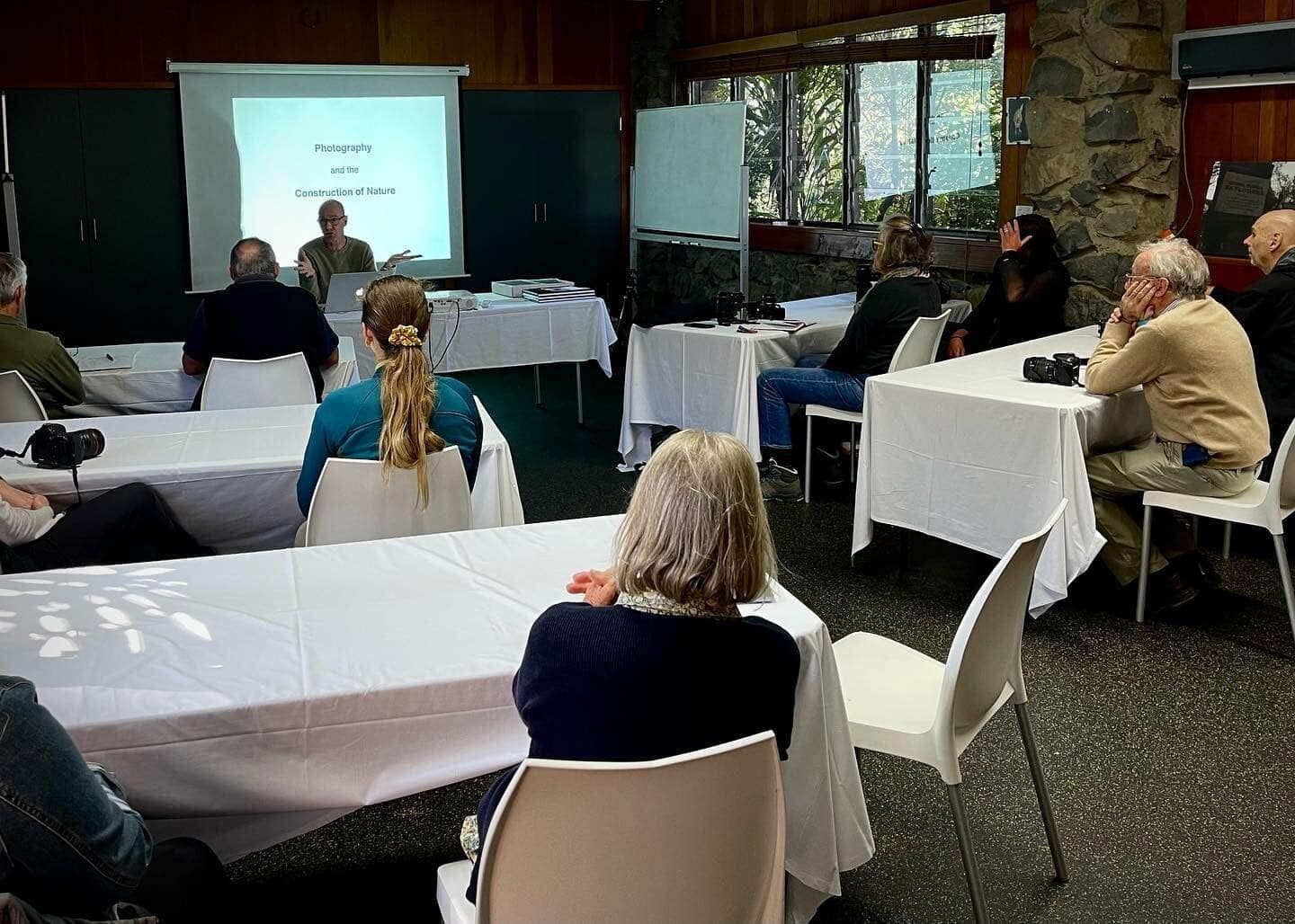 A conference room with round tables, attendees facing a presentation screen, and a stone wall with windows.