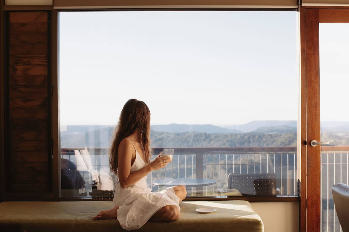 Una mujer sentada en un banco acolchado junto a un ventanal con vistas a montañas.