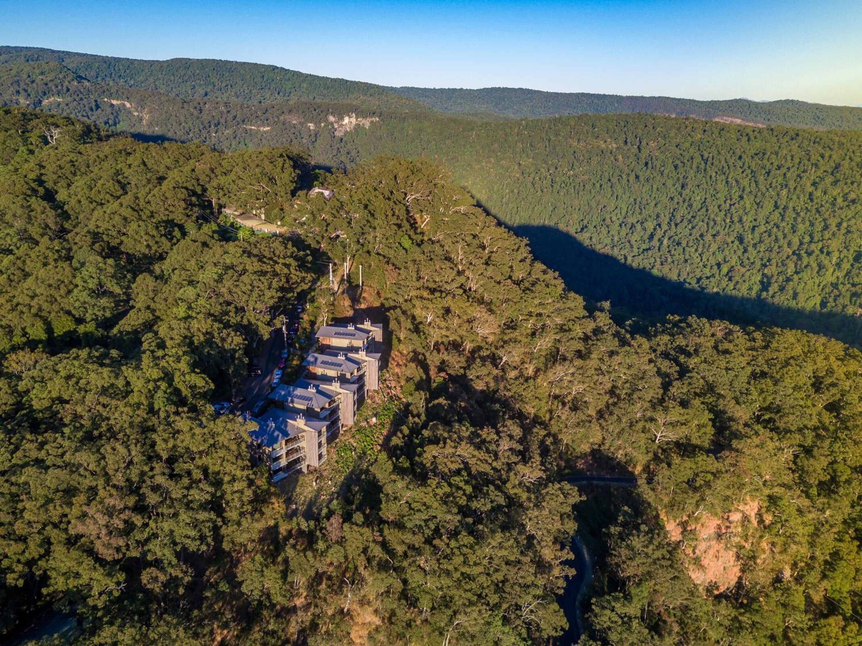 Une vue aérienne d’un complexe hôtelier composé de plusieurs bâtiments nichés au cœur d’une forêt dense, et entouré de collines.