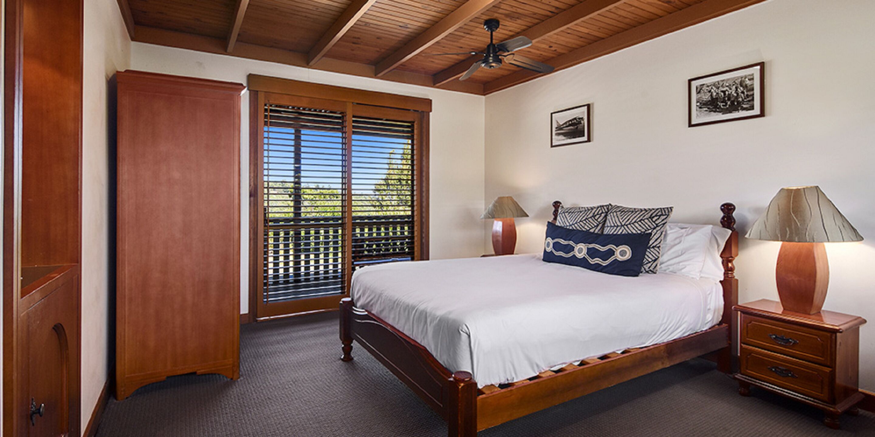 A bedroom with a wooden bed, two bedside tables with lamps, a ceiling fan, a wardrobe, and a window with blinds.
