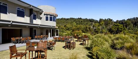 Un edificio con un área de comedor al aire libre techada, varias mesas y sillas, y vistas a una ladera boscosa.