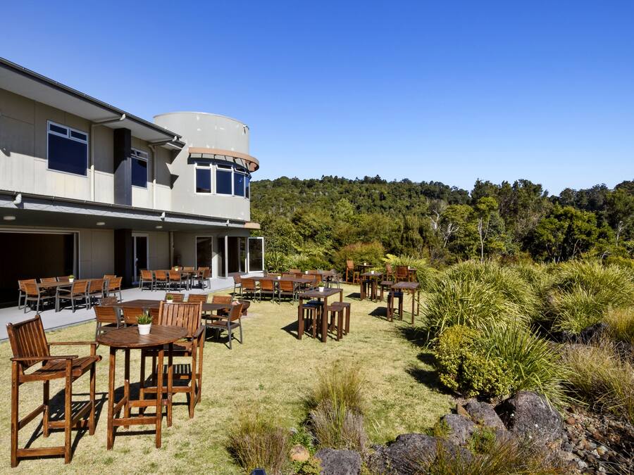 Een gebouw met een overdekt terras, diverse tafels en stoelen, en uitzicht op een beboste heuvel.