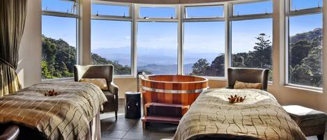 A hotel room with two beds, a wooden tub, and large windows offering a mountain view.