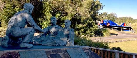 Una estatua de tres figuras sentadas sobre una plataforma de piedra, con un avión azul en el fondo.