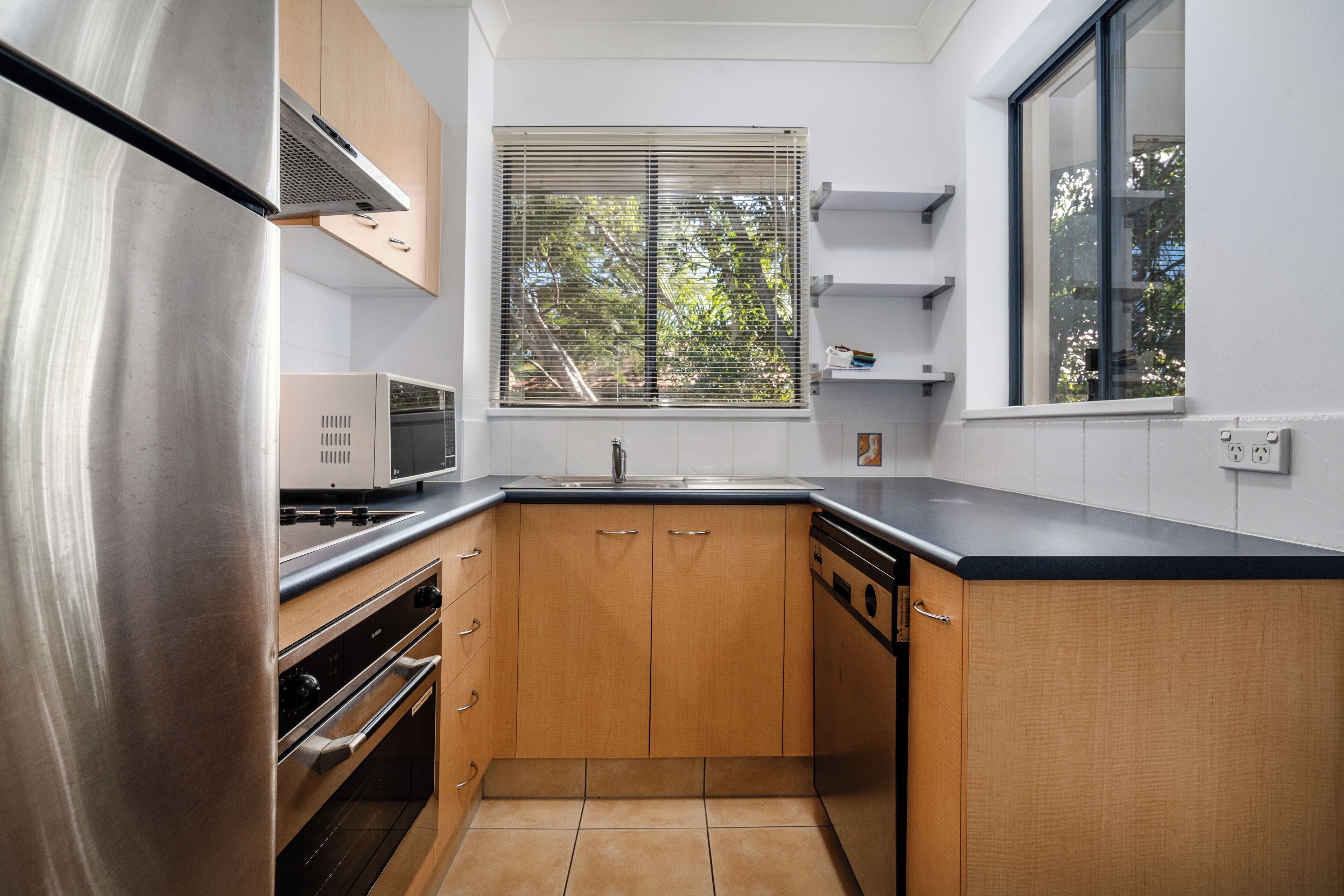 A modern kitchen with stainless steel appliances, wooden cabinets, and a window with blinds.