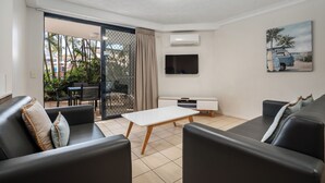 A modern living room with a sofa, a coffee table, and a TV. There is a sliding glass door leading to an outdoor area with plants. - Turtle Beach Resort (Mermaid Beach)