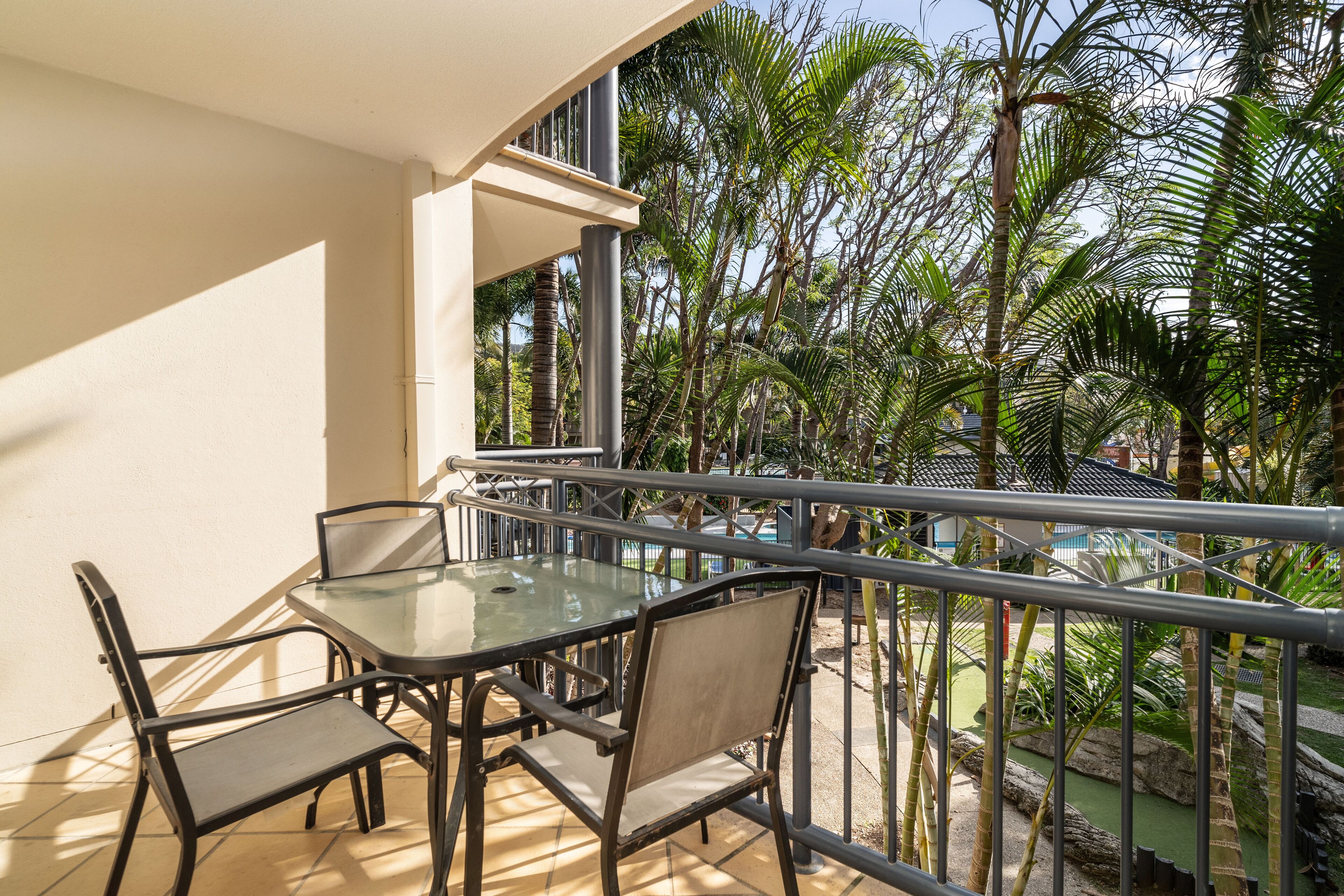 A balcony with a glass table and chairs, overlooking a pool and palm trees.