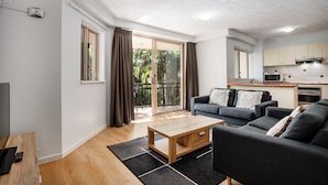 A modern living room with a sofa, coffee table, and a TV. There is a sliding glass door leading to a balcony with a view of greenery. - Turtle Beach Resort (Mermaid Beach)