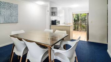 A modern dining area with a glass-top table surrounded by white chairs, a kitchen in the background, and a view of greenery through a glass door.