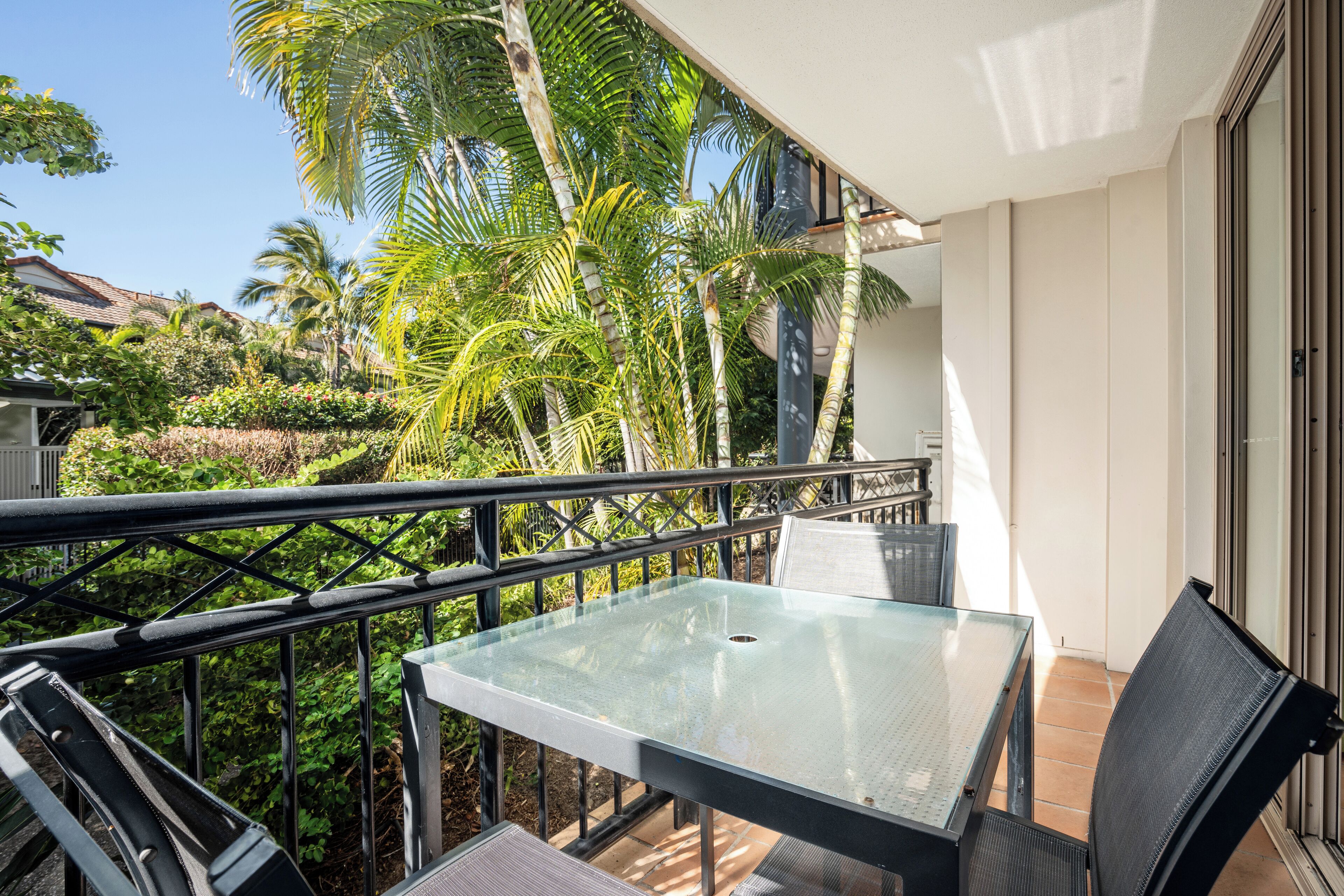 A balcony with a glass table and chairs, surrounded by lush greenery and palm trees.