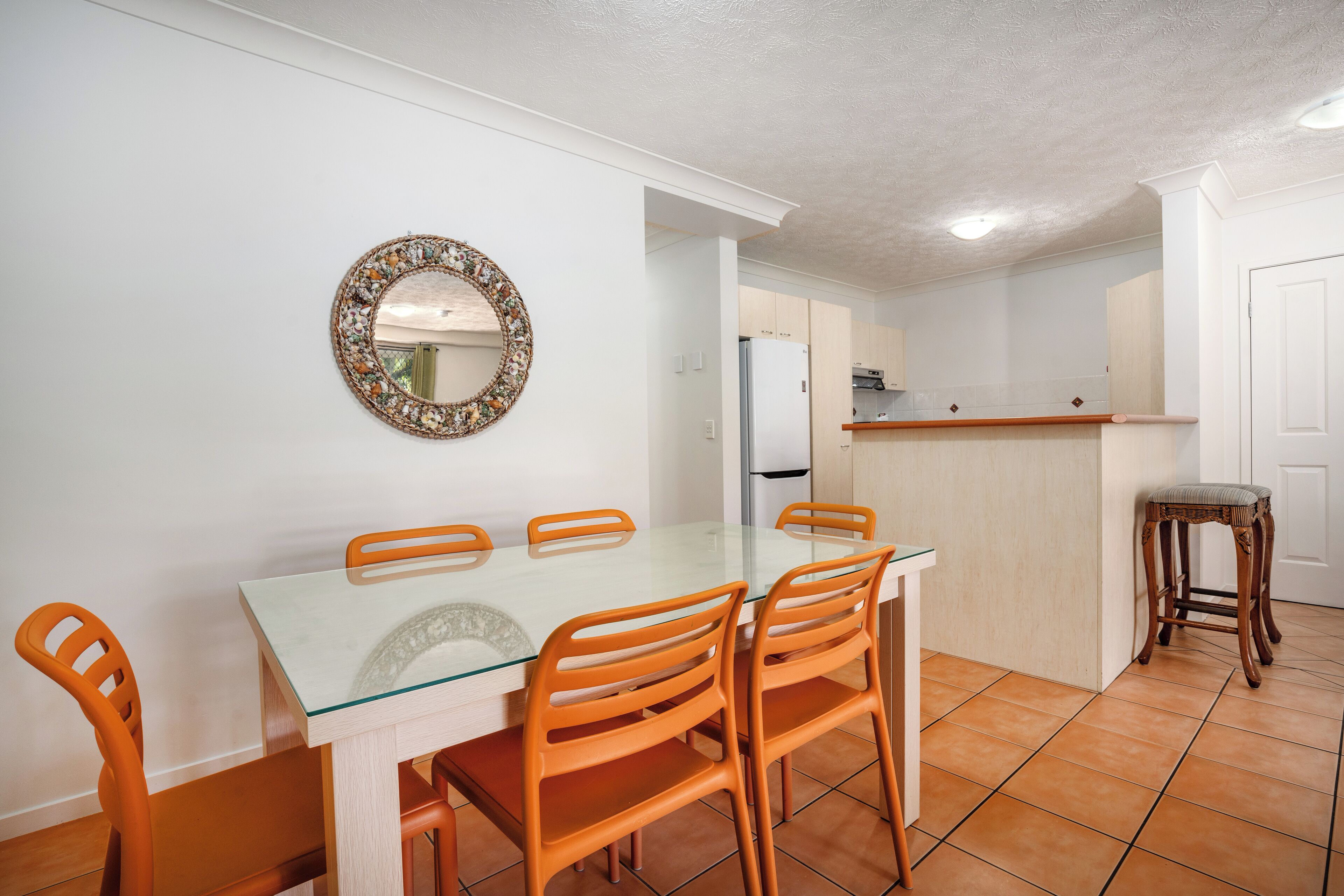 Un coin repas avec une table en verre et des chaises orange, une cuisine en arrière-plan et un tabouret en bois.
