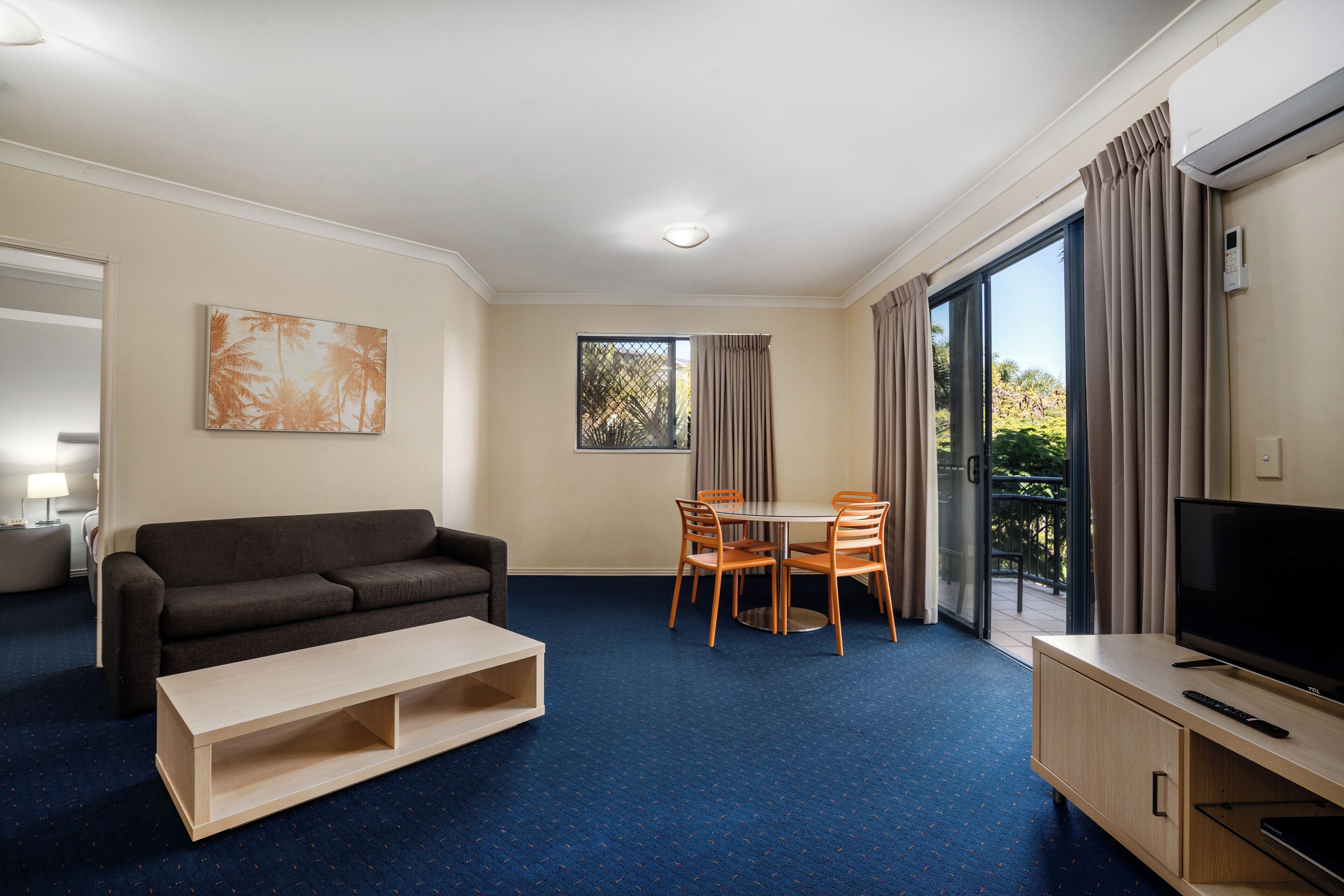 A hotel room with a sofa, a dining table with chairs, a TV, and a large window with curtains.