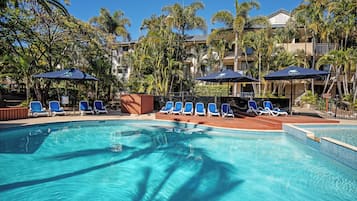 Relax by the resort pool surrounded by lounge chairs and umbrellas, shaded by palm trees and tropical vegetation.