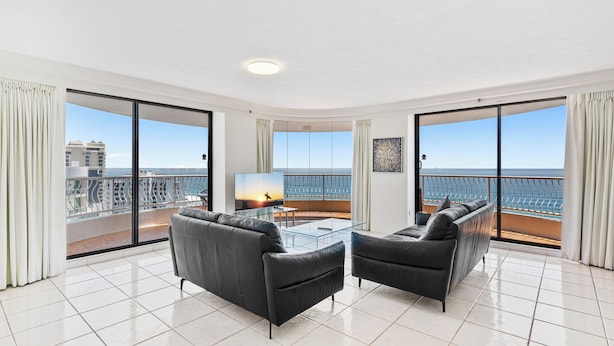 A spacious living room with a glass coffee table, two sofas, and a large window offering a view of the sea and buildings. - Aegean Apartments (Surfers Paradise)