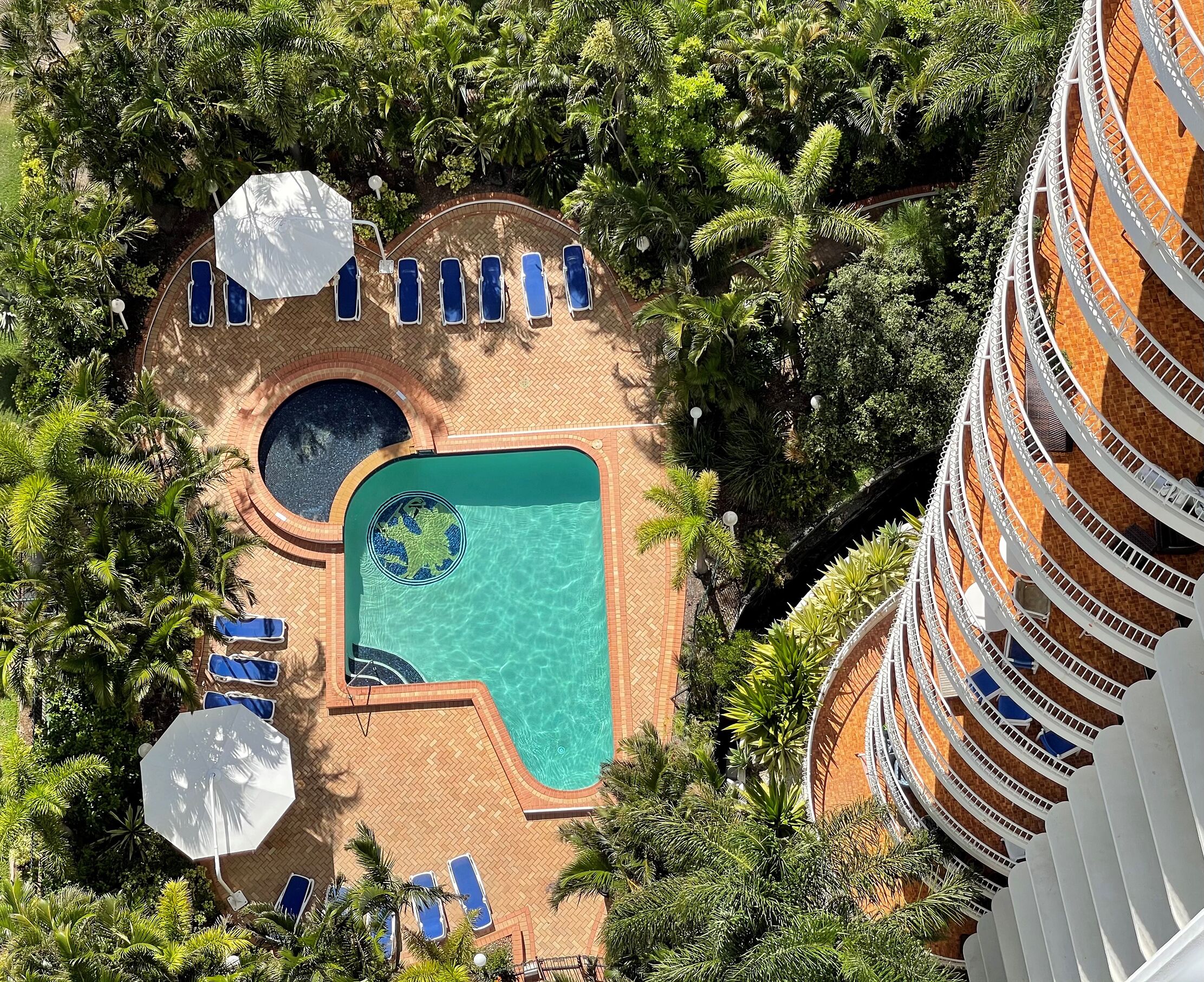 An aerial view of a resort with a swimming pool surrounded by palm trees, lounge chairs, and a circular water feature.