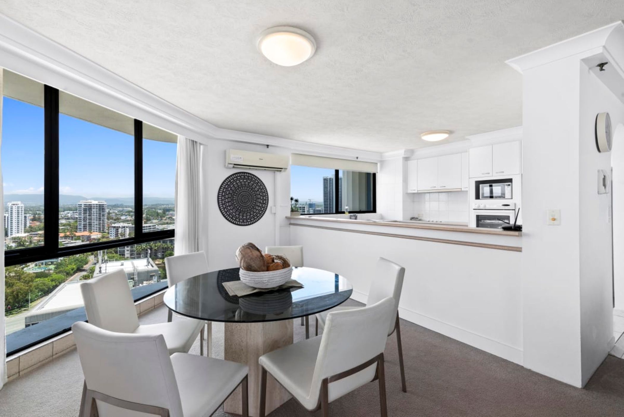 A modern kitchen with white cabinets, a round dining table with white chairs, and a view of the city through large windows.