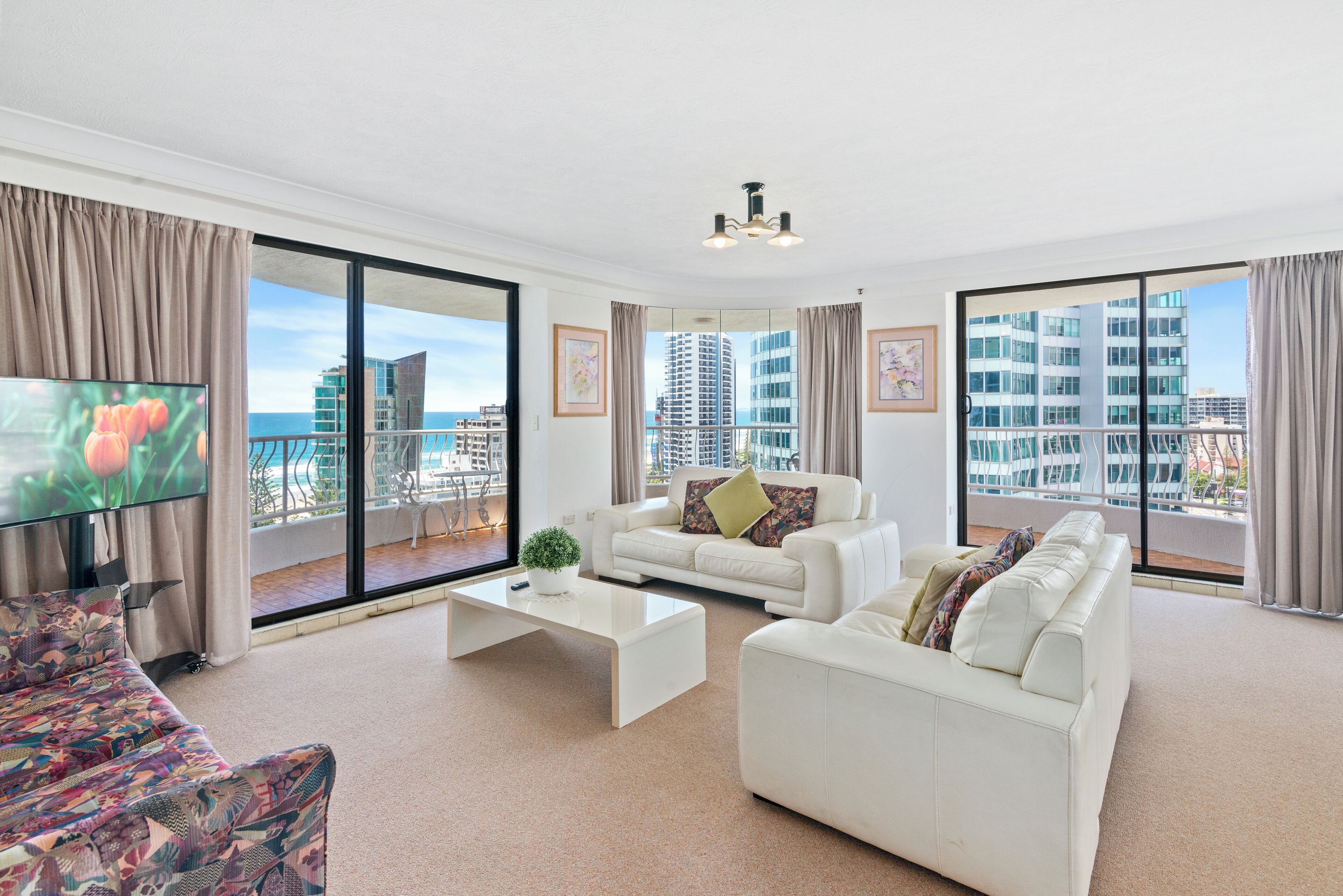 A modern living room with a white sofa set, a flat-screen TV, and large windows offering a city view.