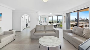 A modern living room with a sofa, a coffee table, and a view of a cityscape through large windows. - Aegean Apartments (Surfers Paradise)