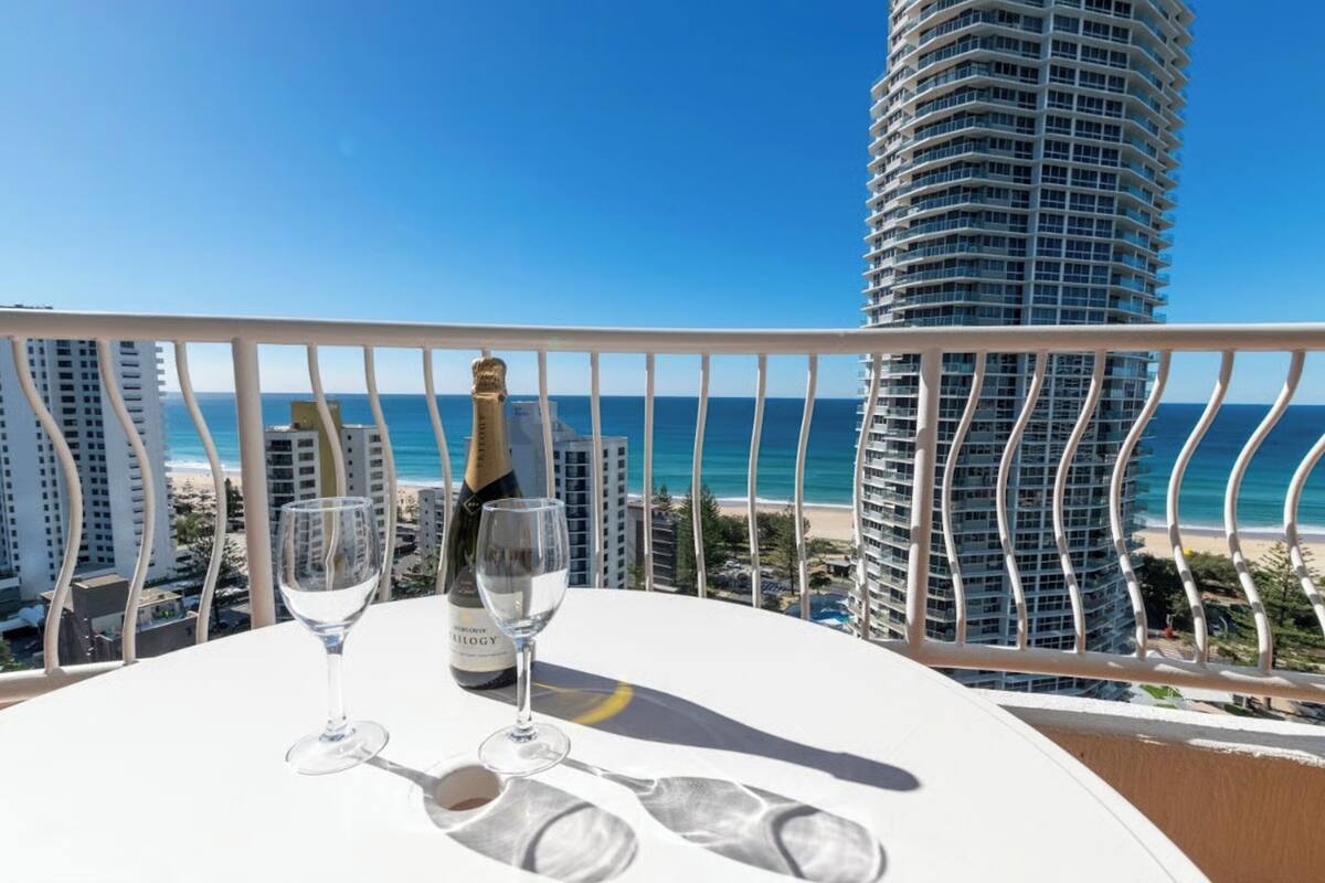 A balcony with a table, a bottle of champagne, and two glasses. The view includes a beach and a tall building.