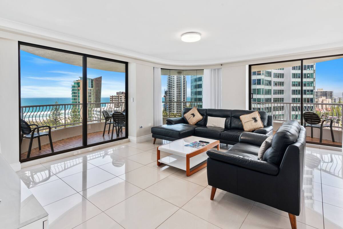 A modern living room with a black leather sofa, a coffee table, and a view of the ocean and high-rise buildings.