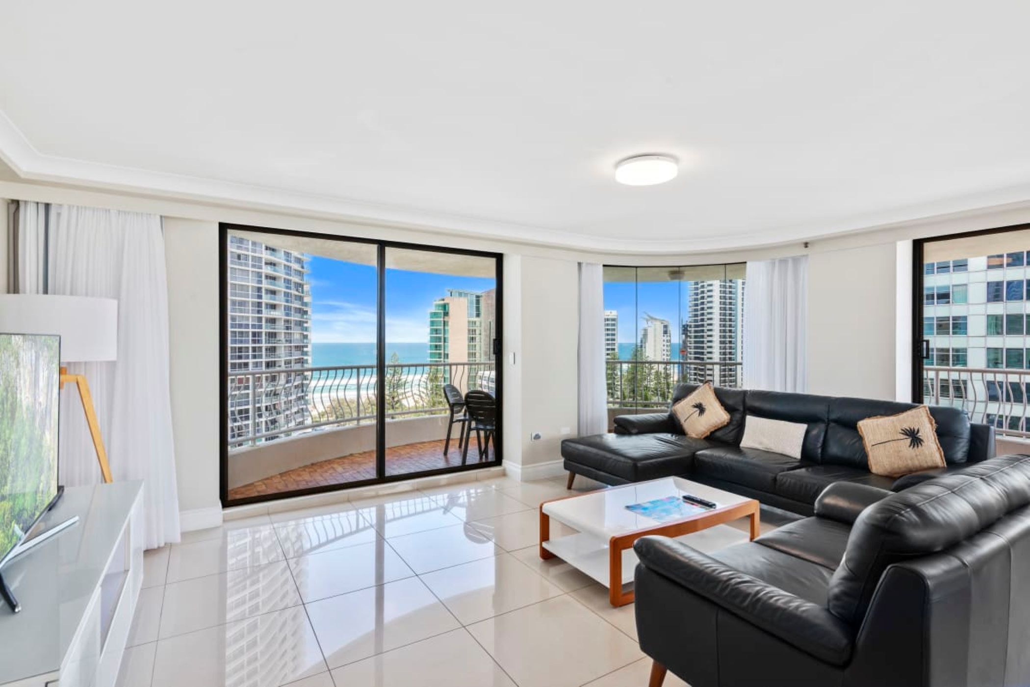 A modern living room with a black leather sofa, a coffee table, and a TV. Large windows offer a view of high-rise buildings and a beach.