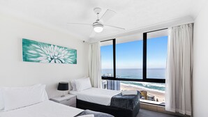 A hotel room with two beds, a ceiling fan, a large window with a view of the beach and ocean, and a painting on the wall. - Aegean Apartments (Surfers Paradise)
