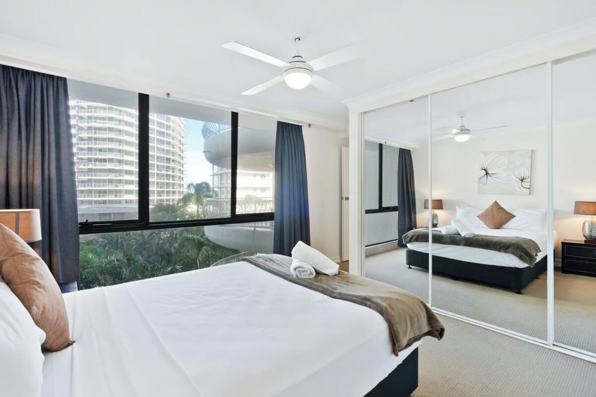 A hotel room with a large bed, a ceiling fan, a view of a cityscape, and a sliding glass door.