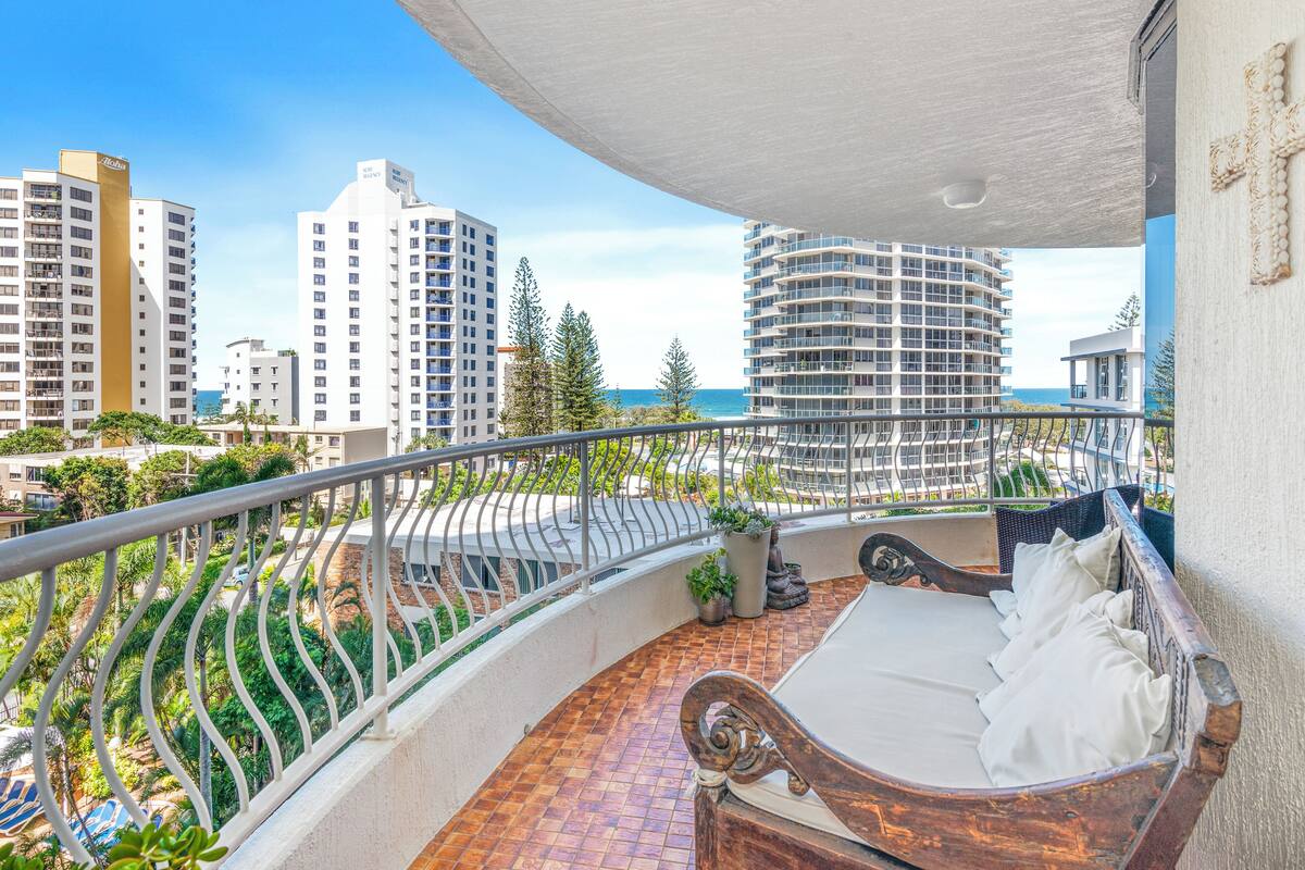 A balcony with a bed, potted plants, and a view of tall buildings and the ocean.