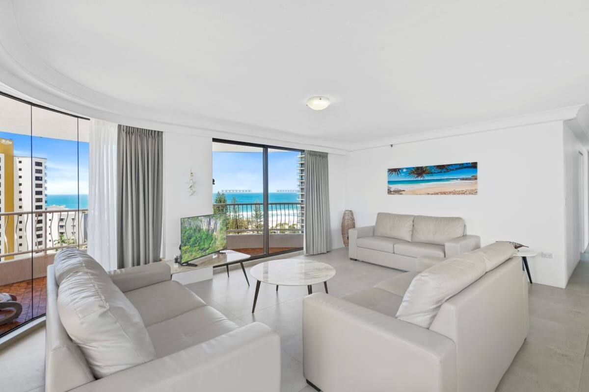 A modern living room with a large window, a sofa set, a coffee table, and a painting of a beach scene.