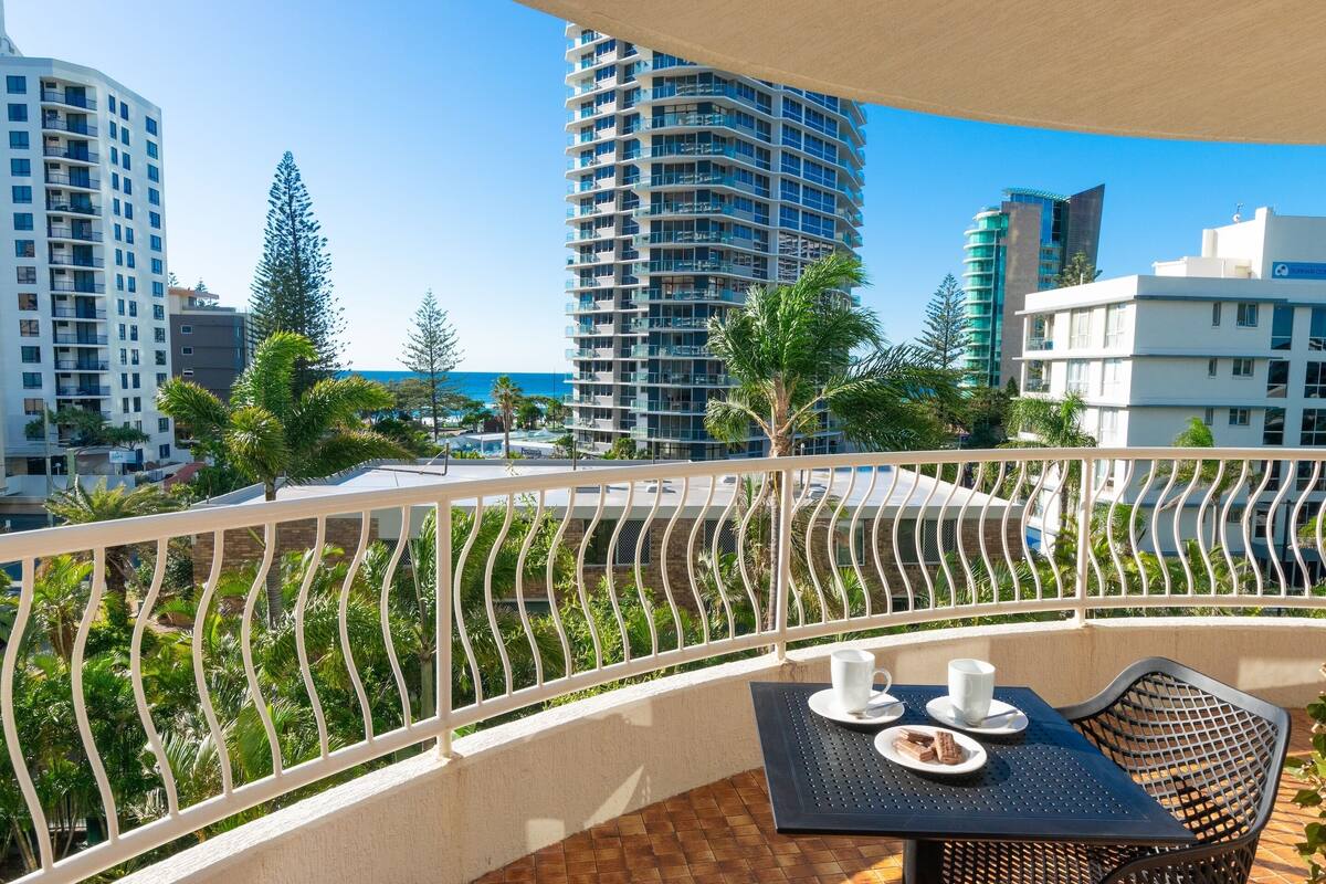 A balcony with a table and chairs, overlooking a cityscape with tall buildings and a clear sky.