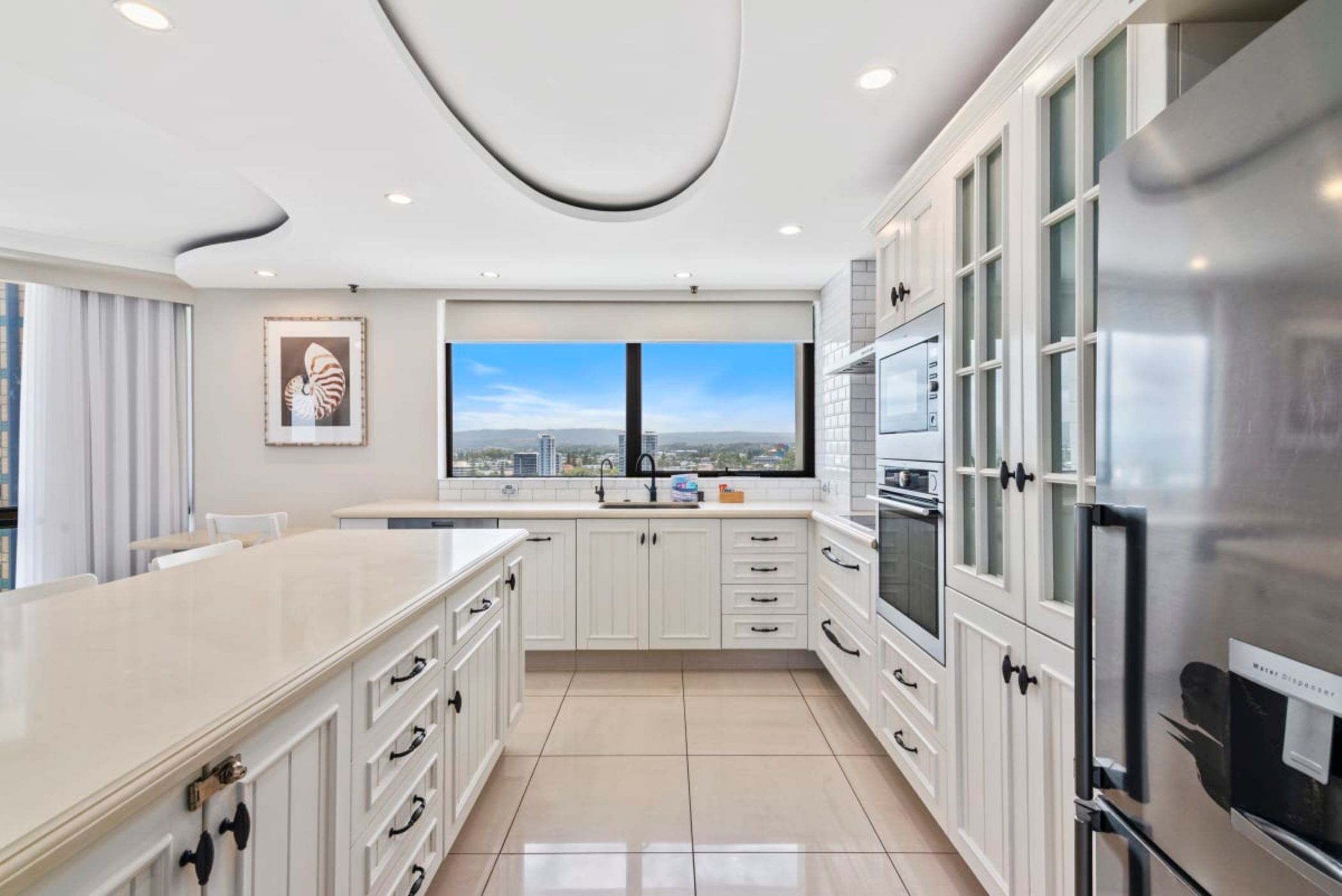 A modern kitchen with white cabinetry, a large island, and a view of the city through a window.