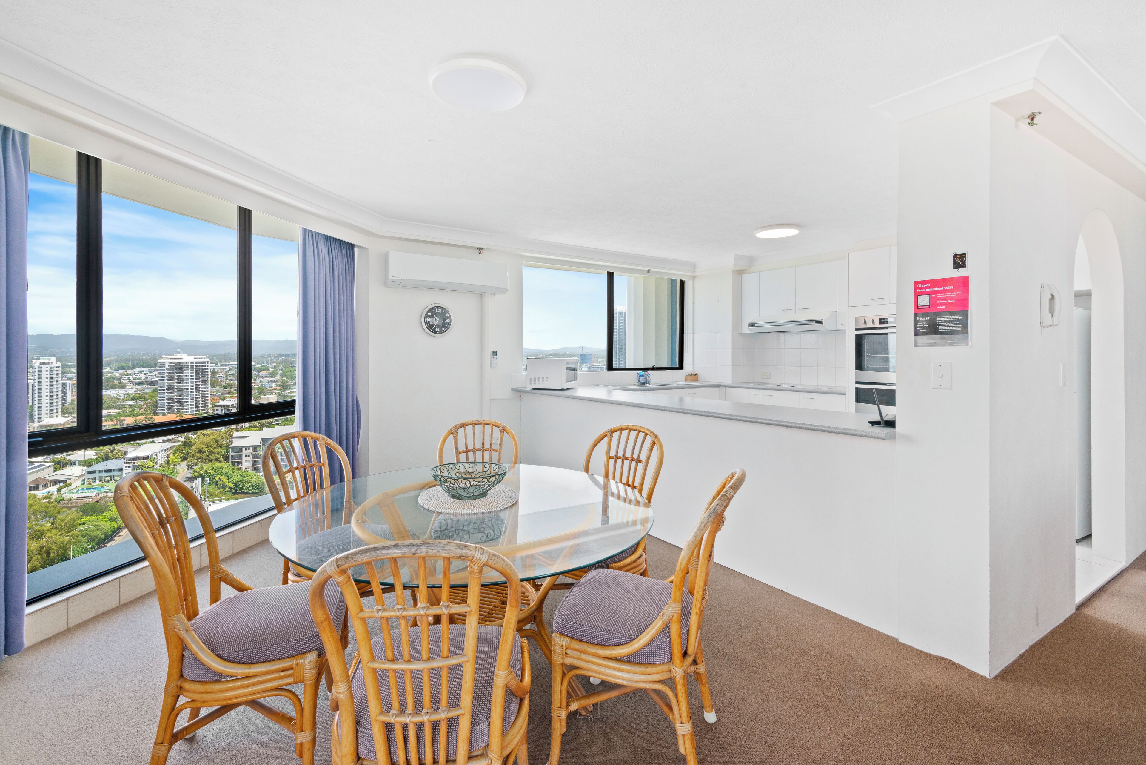 A modern kitchen with white cabinets, a dining area with wicker chairs and a glass table, and a city view through large windows.