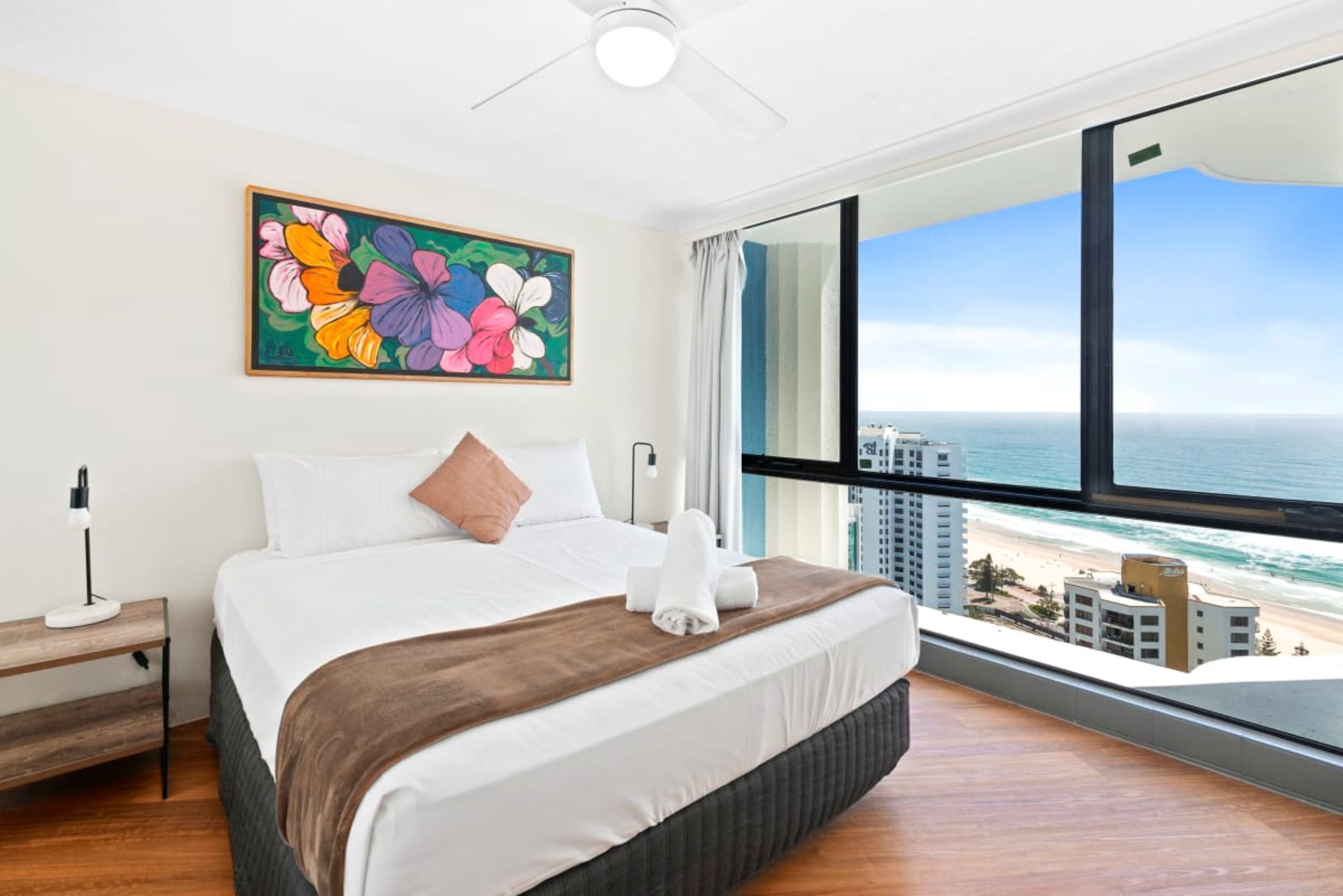 A hotel room with a large bed, a painting of flowers, a view of the ocean, and a cityscape.
