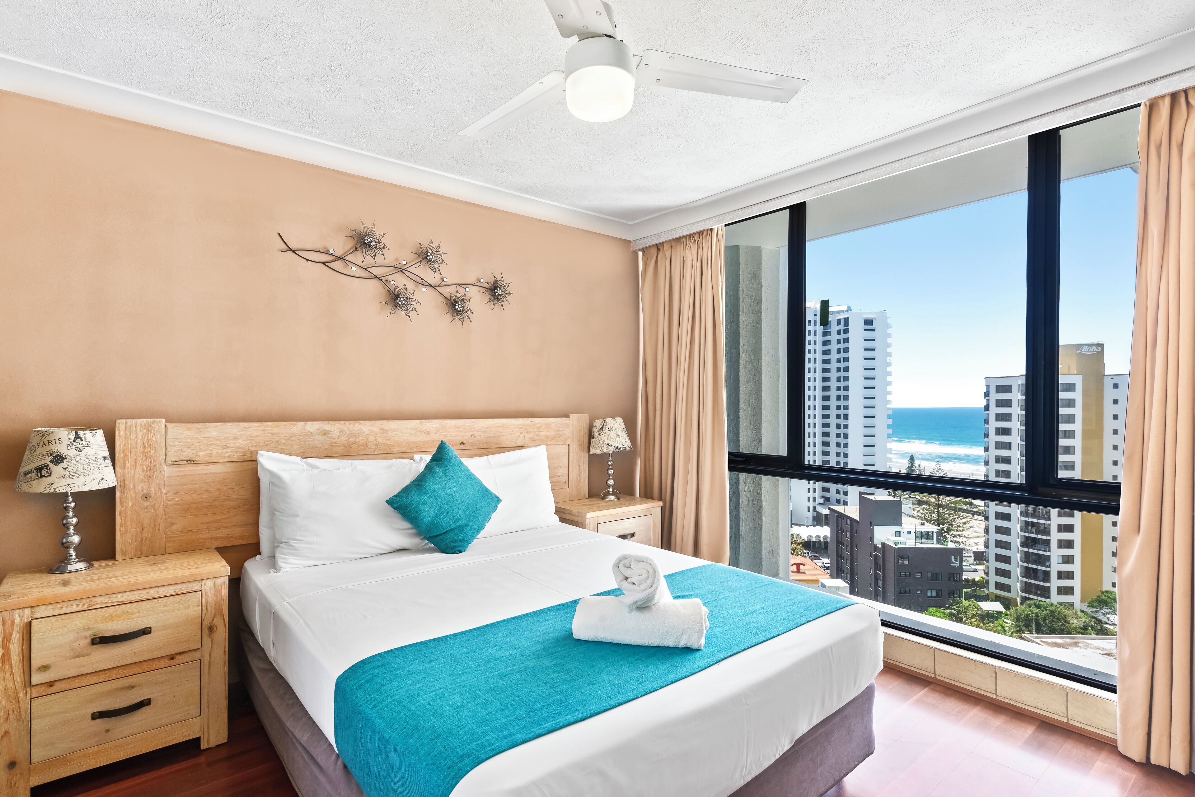 A hotel room with a large bed, two bedside tables, a ceiling fan, and a view of the city and ocean.