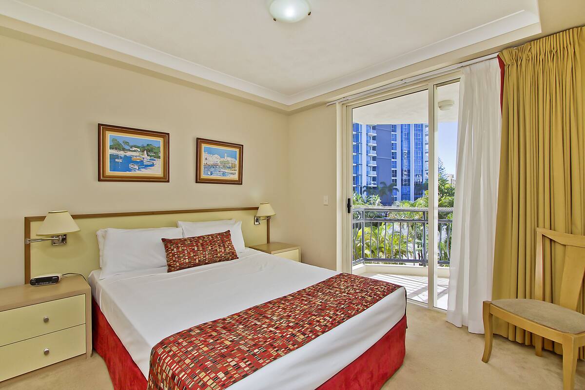 Una habitación de hotel con una cama, dos cuadros en la pared, una mesita de noche, una silla y un balcón con vistas a edificios.